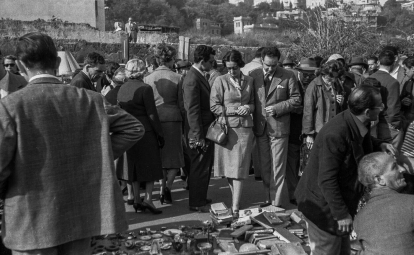 A black and white photo of people at an outdoor market with mid-century clothing, browsing items on the ground; buildings in back.