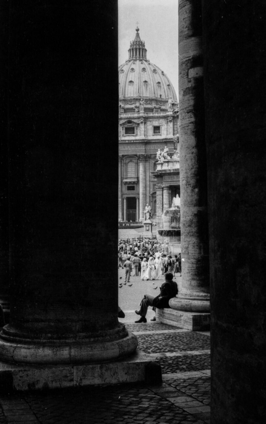 Una persona su una panchina tra due colonne di pietra; sullo sfondo la folla, la fontana e la cupola della Basilica di San Pietro.