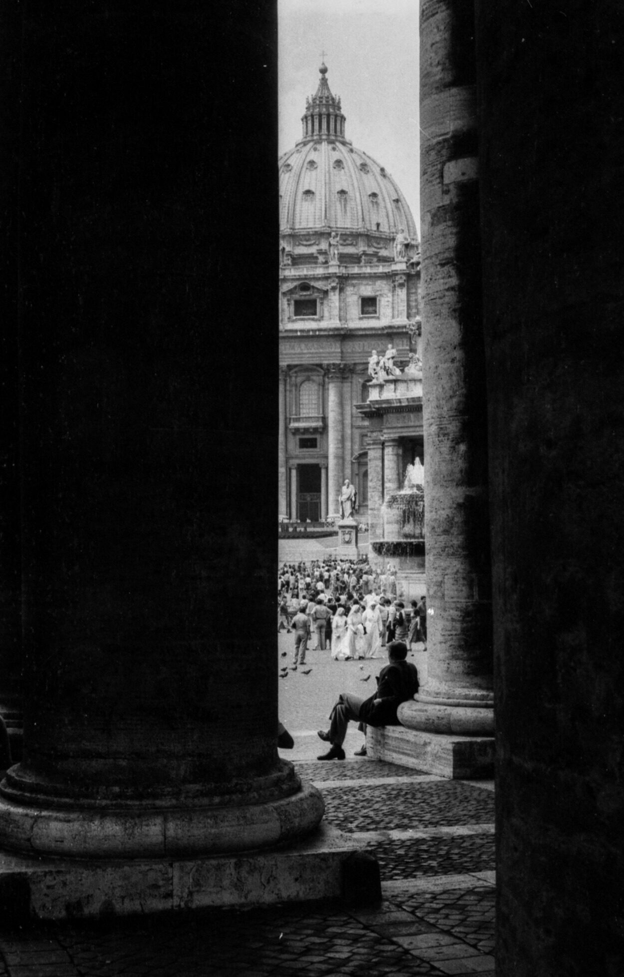 St. Peter’s Basilica - Black and white photo of the dome framed by columns; person on bench, crowd near fountain in bright square.
