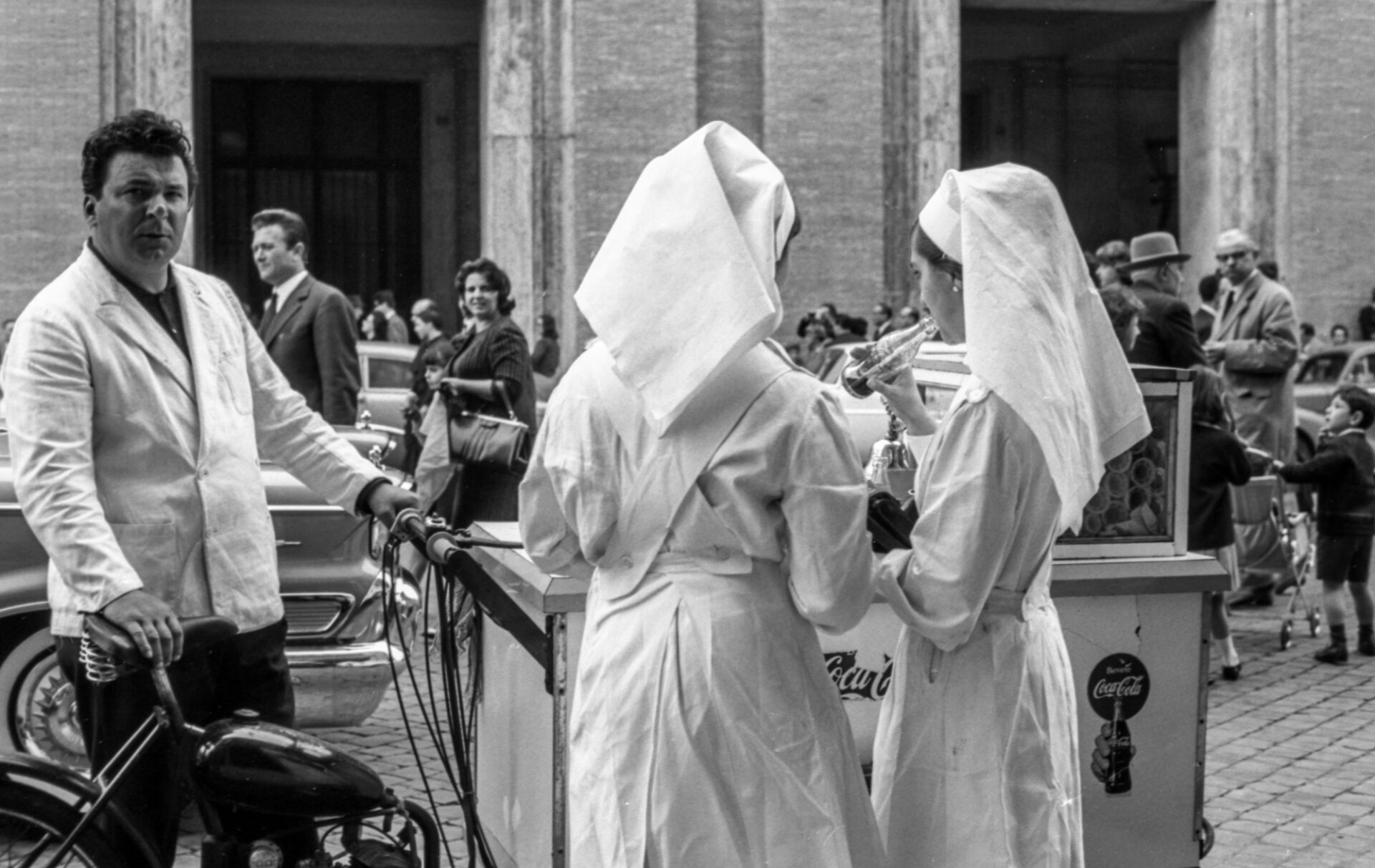 A black-and-white photo: Two women in nurse uniforms buy snacks from a street vendor on a cobblestone street with cars and people.