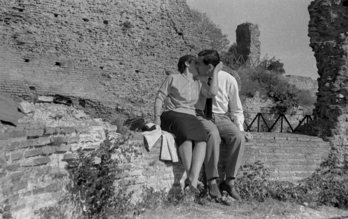 A black and white photo of a mid-century dressed couple kissing on a stone wall with ancient ruins and overgrown grass behind them.