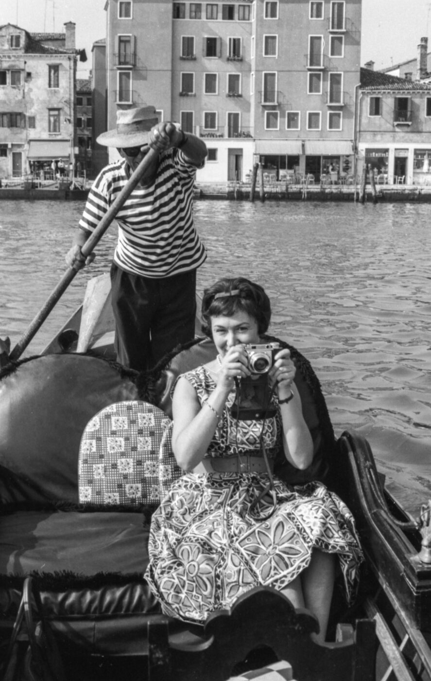 A smiling woman in a patterned dress sits on a gondola with camera; behind her, a striped gondolier rows past buildings along the canal.