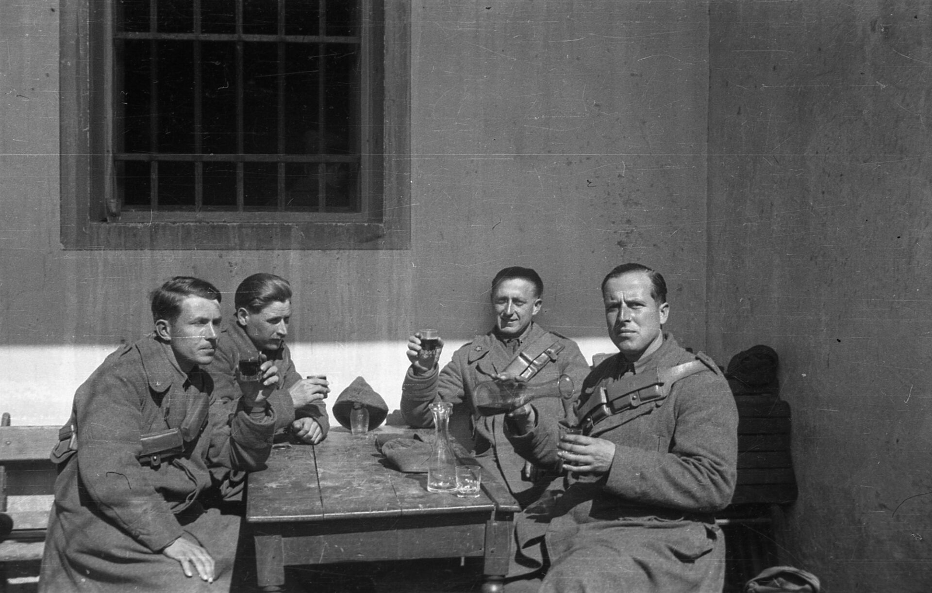 Four soldiers in uniform sit at a wooden outdoor table, drinks in hand, facing the camera by a barred window and plain wall.
