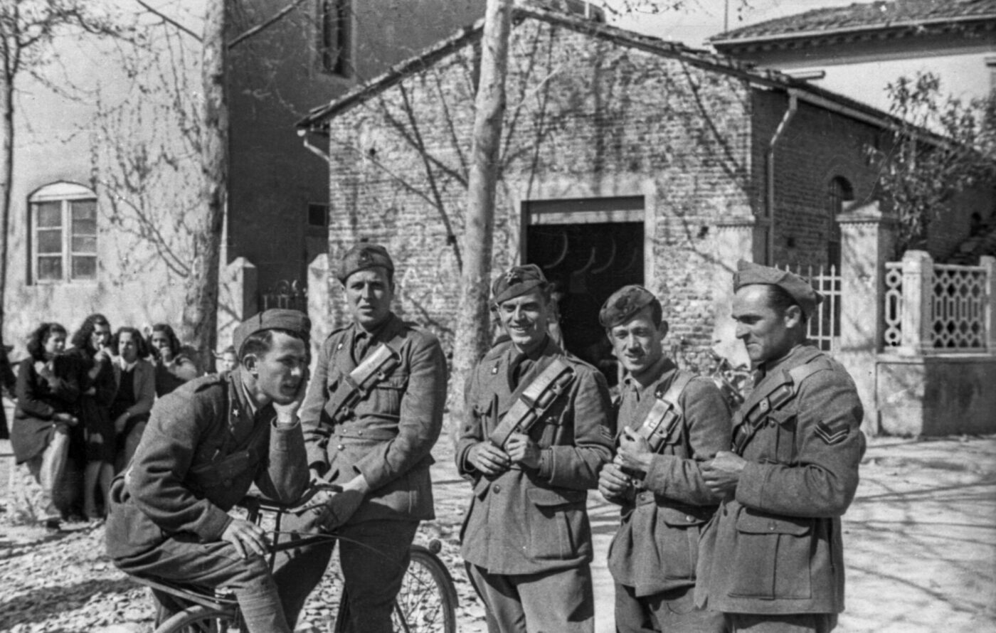 Five soldiers in uniform, some with rifles, pose by a brick building; one on a bicycle. Women and trees appear in the shadowy background.
