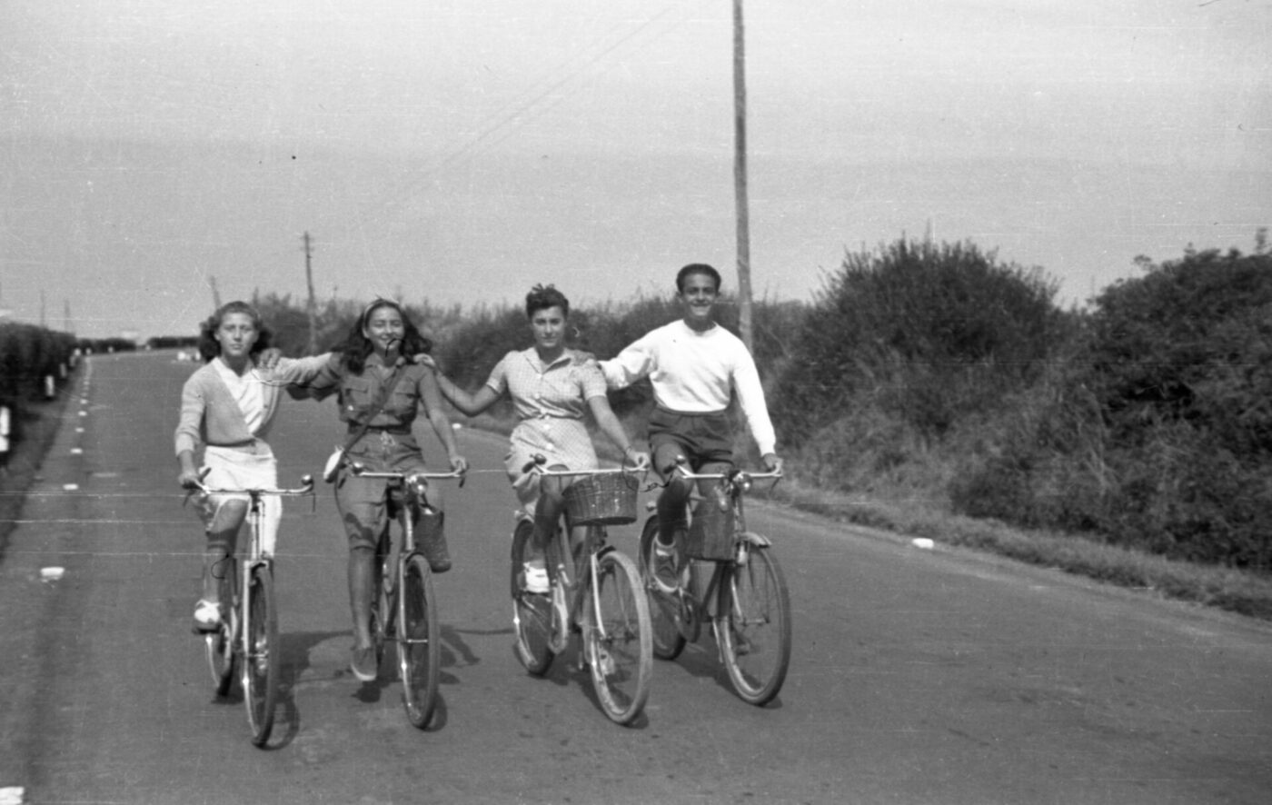 Four people ride bikes side by side on a rural road, arms linked and smiling. Black-and-white scene with bushes and utility poles behind.
