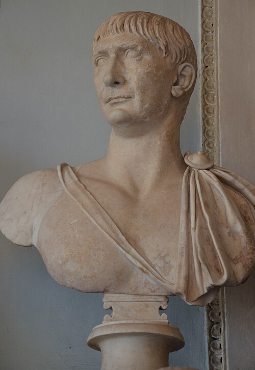 Marble bust of a man with short hair and draped clothing on a pedestal before a plain wall with trim; classical, ancient style.