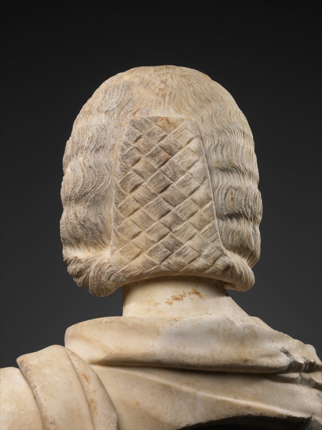 Marble sculpture—head and shoulders from behind, ornate lattice wig, wavy hair, draped clothing; dark backdrop highlights details.