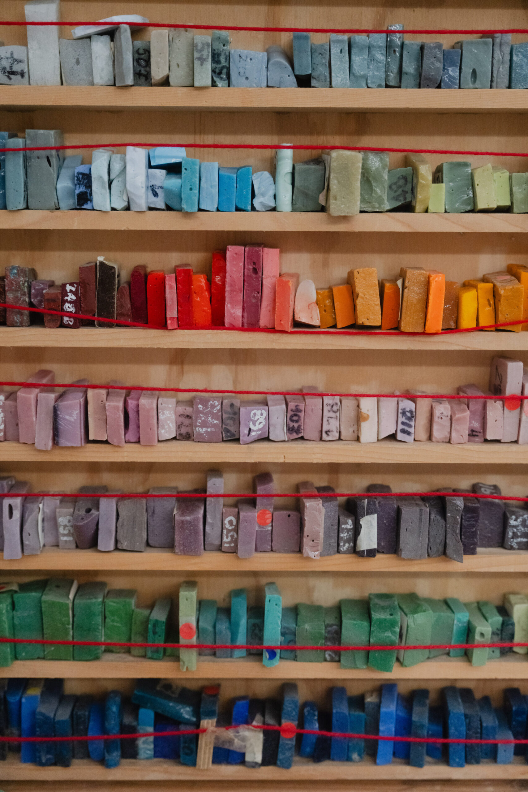 Uno scaffale di legno contiene file di campioni di argilla colorata, ordinati per tonalità, ciascuno etichettato con numeri o codici.
