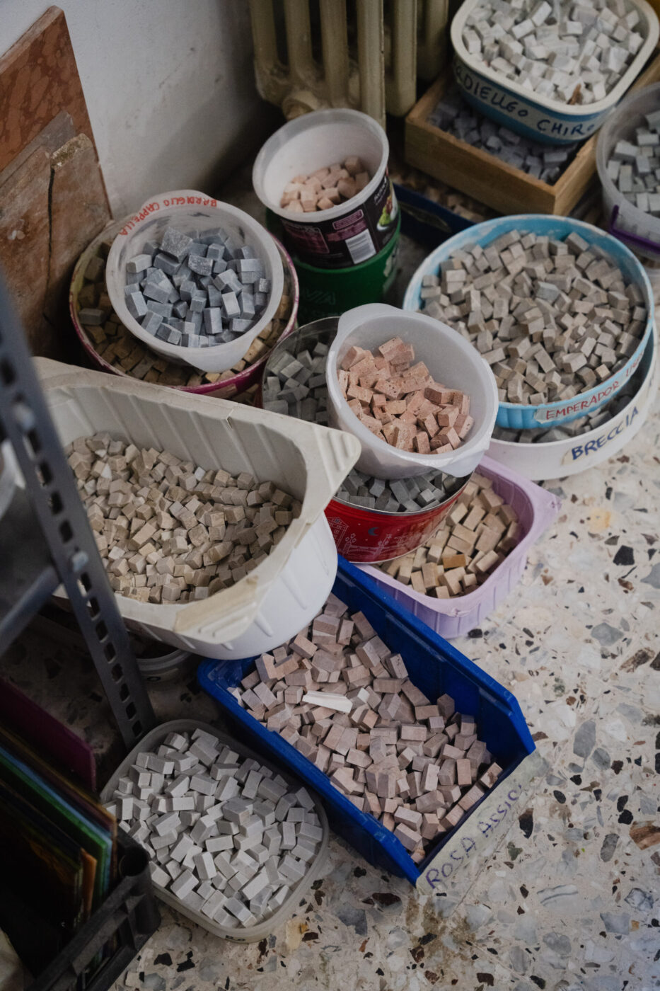 Contenitori assortiti di tessere quadrate di mosaico rosa, grigie e bianche si trovano sul pavimento macchiato, accanto a una mensola di metallo e a un termosifone.