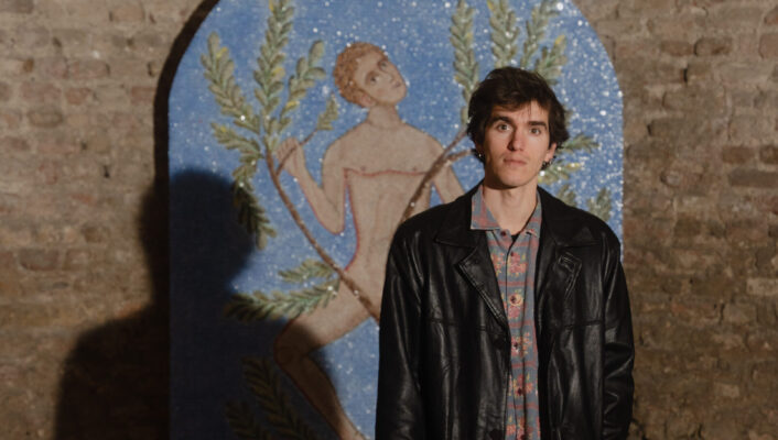 Ismaele Nones with his work "Verso l’Alto, verso la Terra: Sguardo al firmamento"A person in a black leather jacket stands before a brick wall with a vivid mosaic and warm light casting shadows behind them.