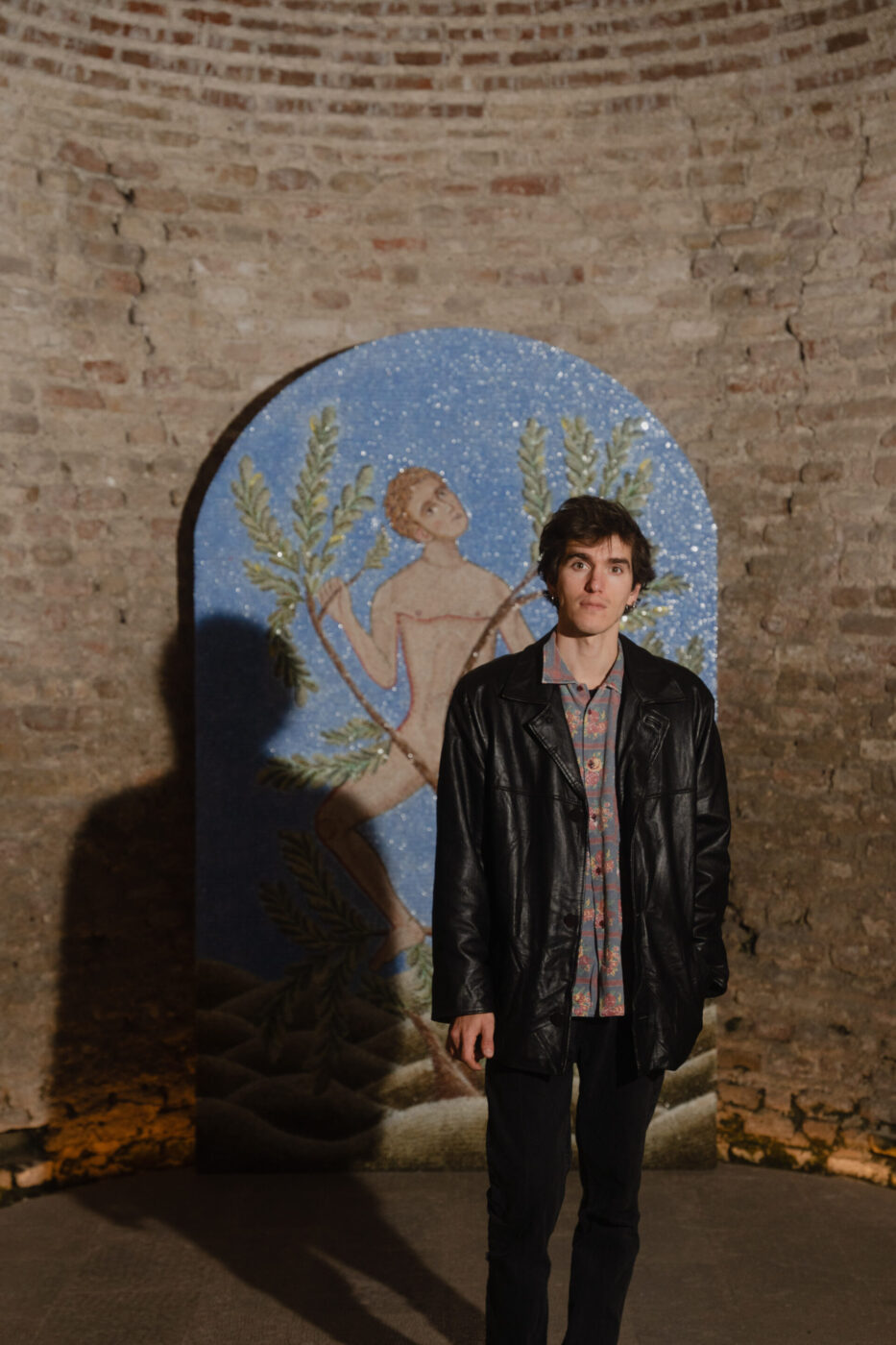 Una persona con una giacca di pelle nera si trova all'interno di un muro di mattoni e di un vivace mosaico che mostra una figura nuda che tiene in mano dei rami verdi.