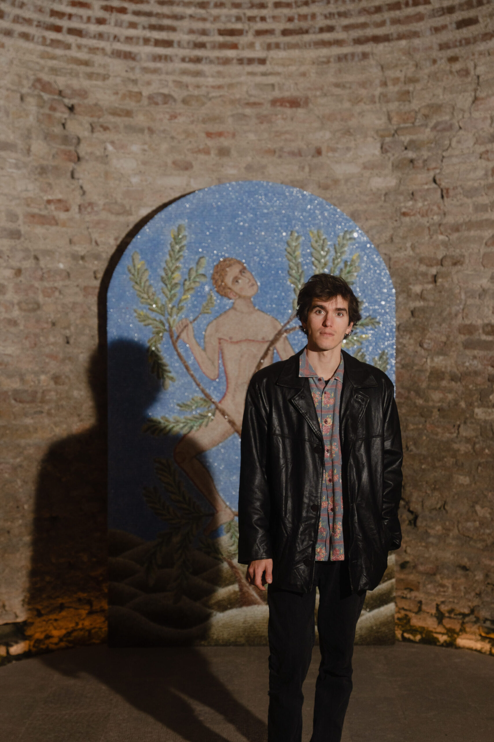 Una persona con una giacca di pelle nera si trova all'interno di un muro di mattoni e di un vivace mosaico che mostra una figura nuda che tiene in mano dei rami verdi.