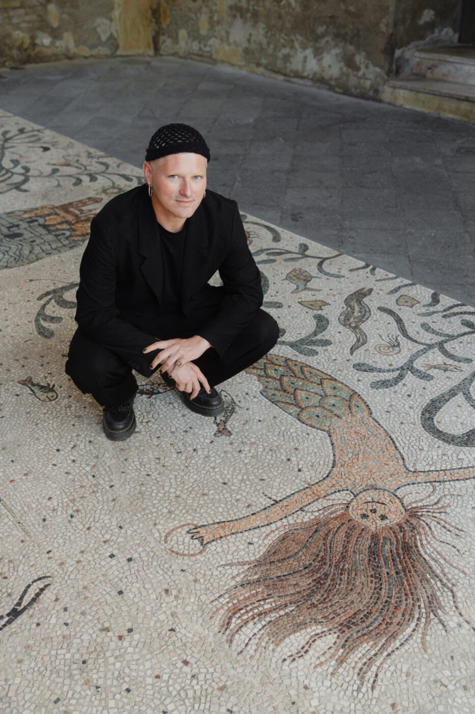 Una persona in abito nero e cappello si accovaccia su un pavimento a mosaico con sirene colorate; sullo sfondo un muro di pietra strutturato.