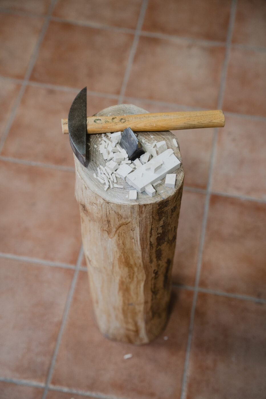 Un ceppo di legno e un'accetta poggiano su frammenti di piastrelle su un pavimento piastrellato. Piccoli frammenti bianchi creano un effetto di dispersione e rottura intorno a loro.