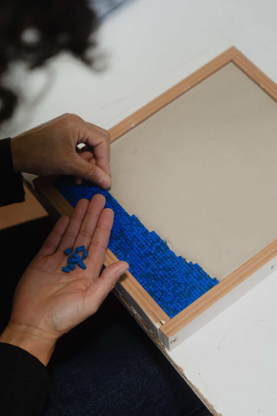 Una persona dispone piccole tessere di mosaico blu in file ordinate su una superficie beige all'interno di una cornice di legno, tenendo altre tessere nell'altra mano.