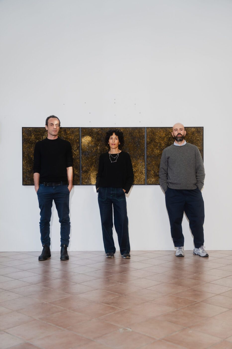 : Tre persone distanziate da una parete bianca con tre dipinti astratti in oro e nero, indossano abiti scuri e casual.