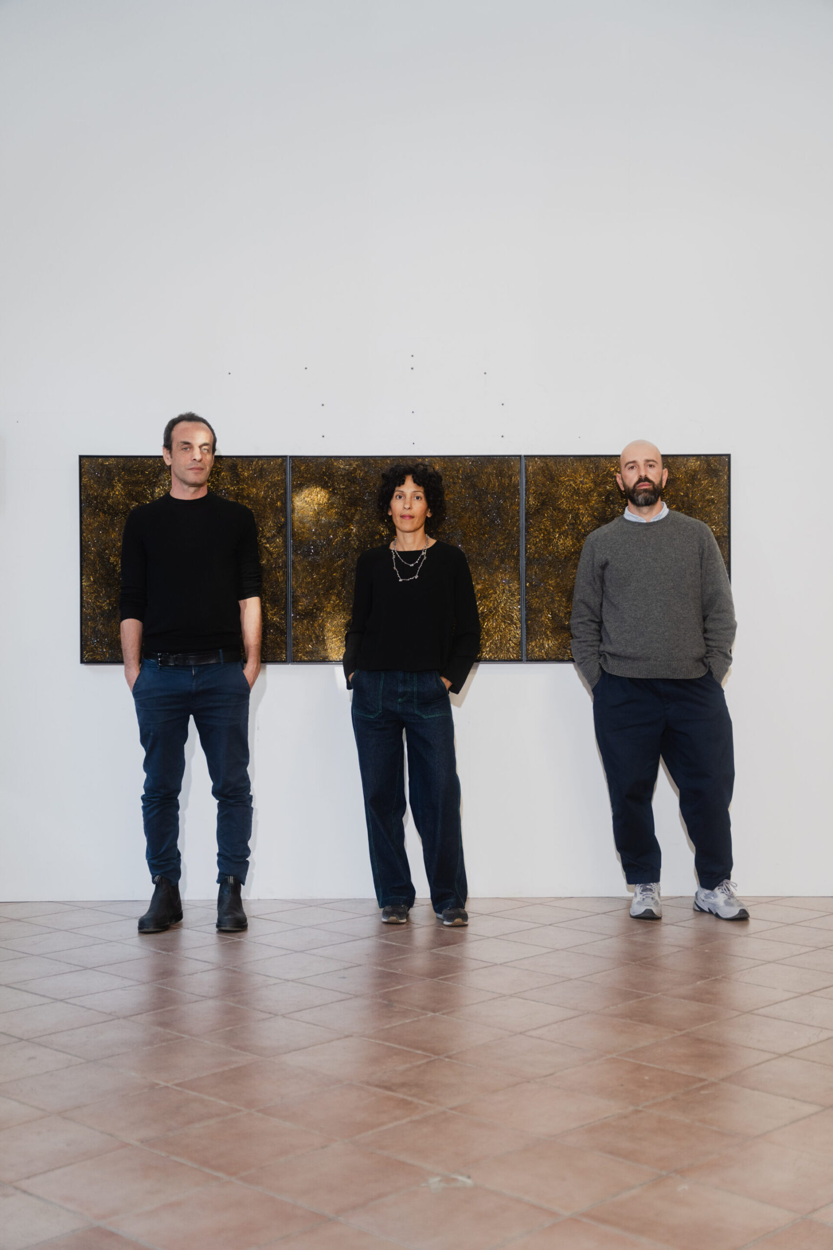 : Tre persone distanziate da una parete bianca con tre dipinti astratti in oro e nero, indossano abiti scuri e casual.