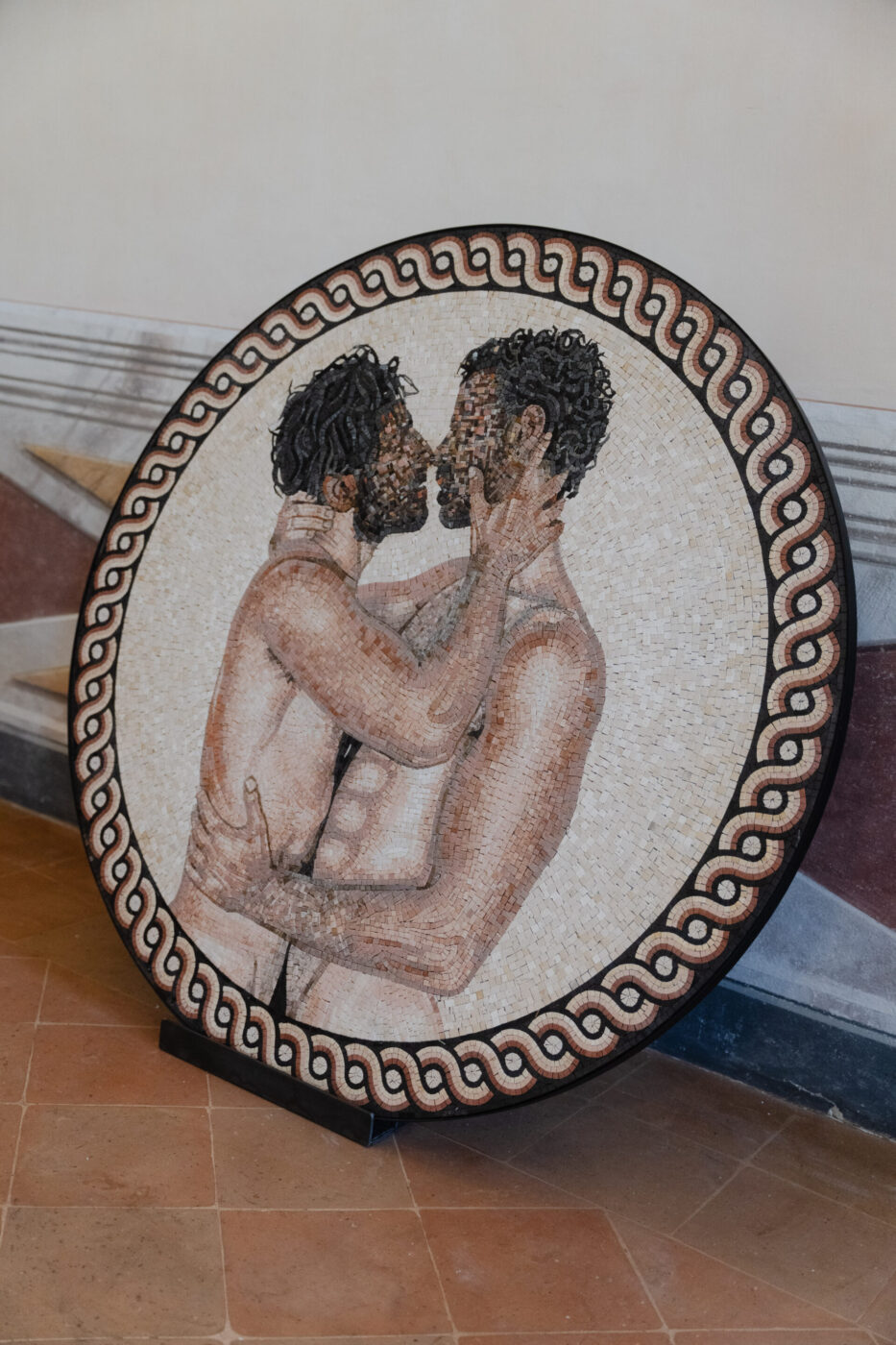Un mosaico circolare di due uomini che si abbracciano e si baciano in toni caldi, con un bordo ondulato in bianco e nero, poggia su un pavimento piastrellato.