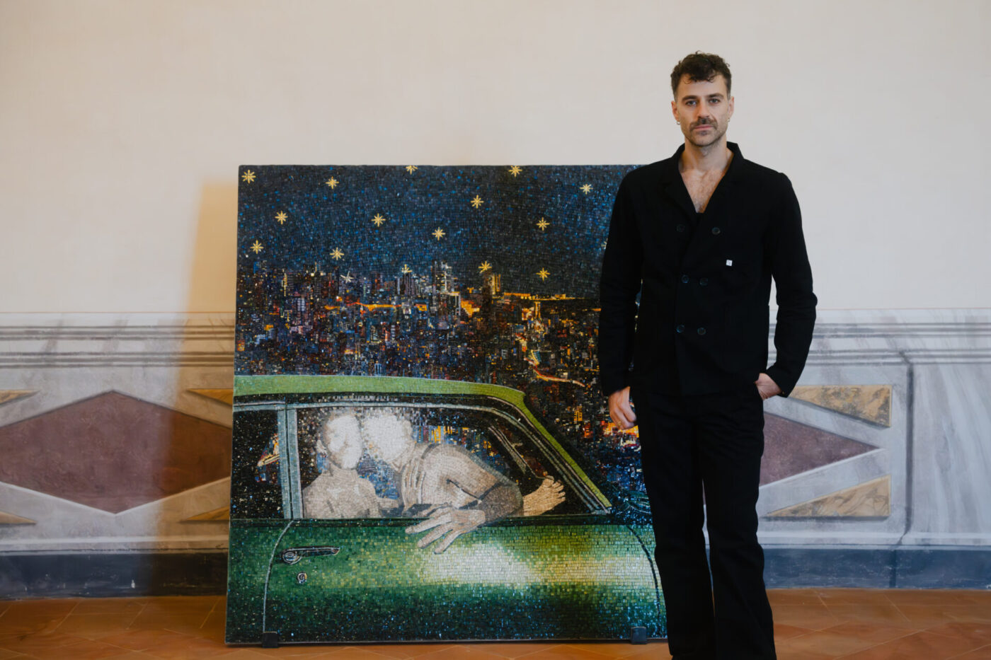 Un uomo vestito di nero si trova al chiuso accanto a un grande dipinto di due persone che si baciano in un'auto verde, con alle spalle un paesaggio urbano e un cielo stellato.