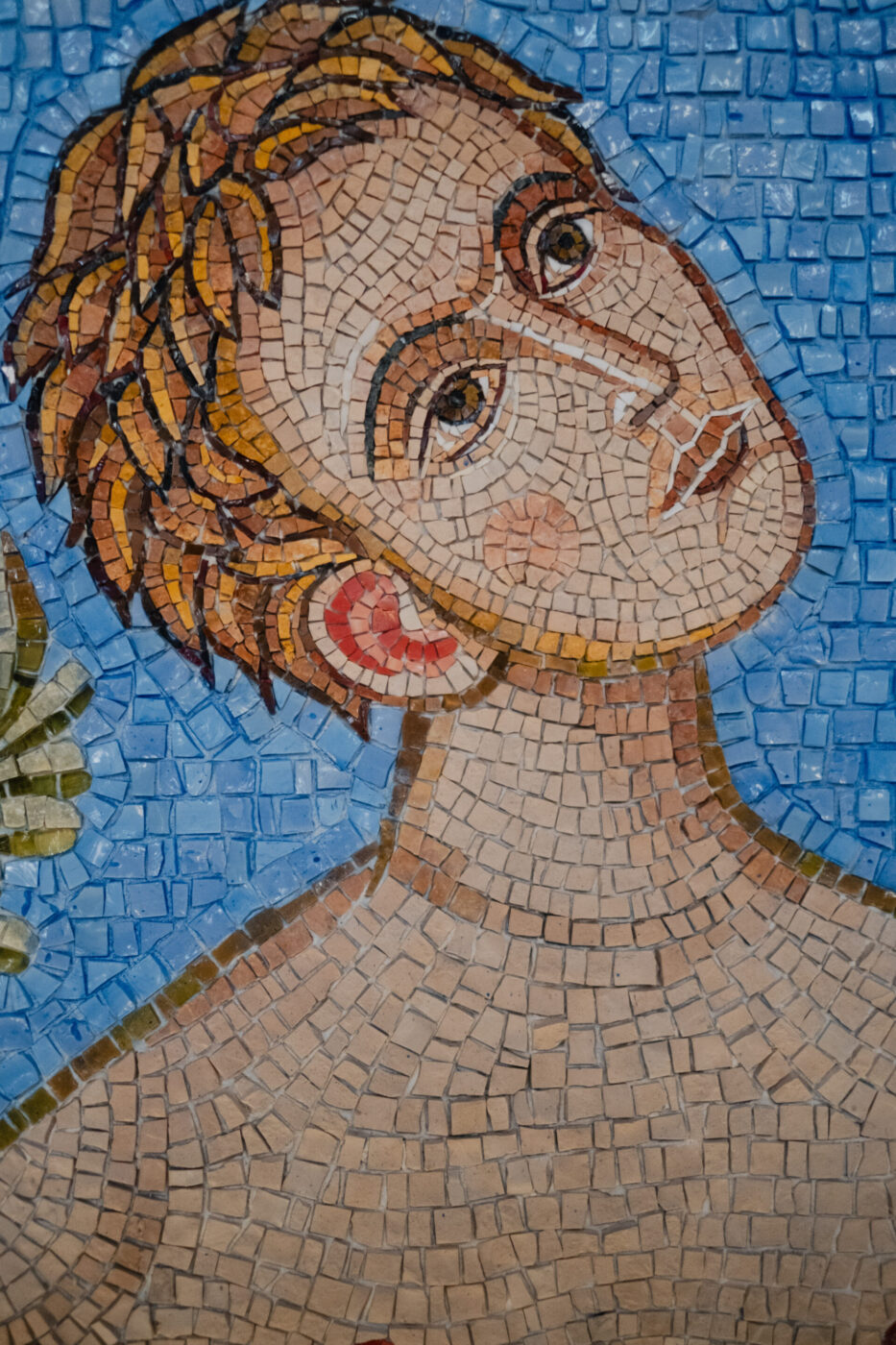Opera d'arte a mosaico raffigurante una persona con capelli castano chiaro, spalle scoperte, che guarda verso l'alto su uno sfondo di piastrelle blu.