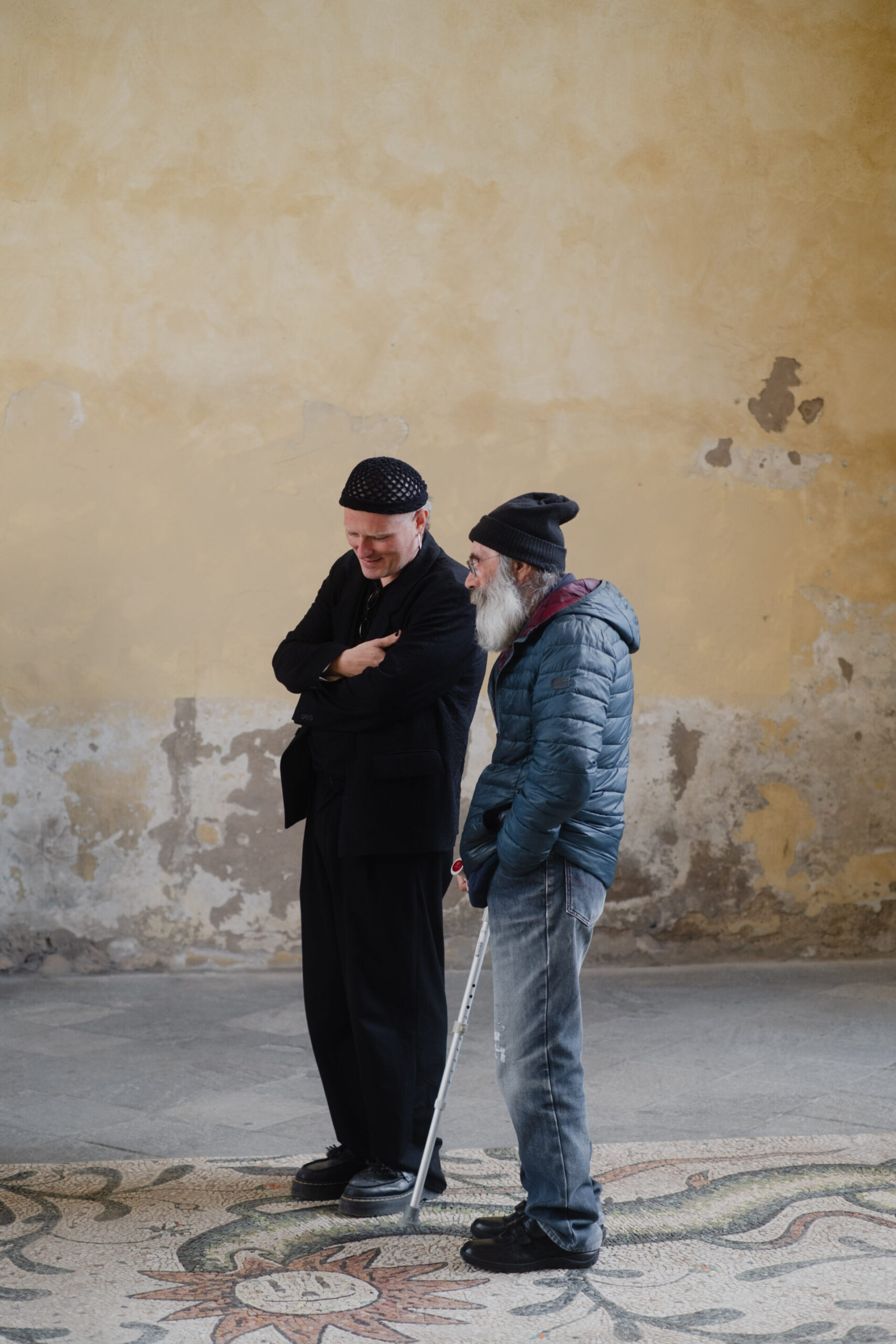Due uomini anziani chiacchierano davanti a una parete strutturata; uno sorride in abito nero, l'altro si appoggia a un bastone in giacca e berretto blu.