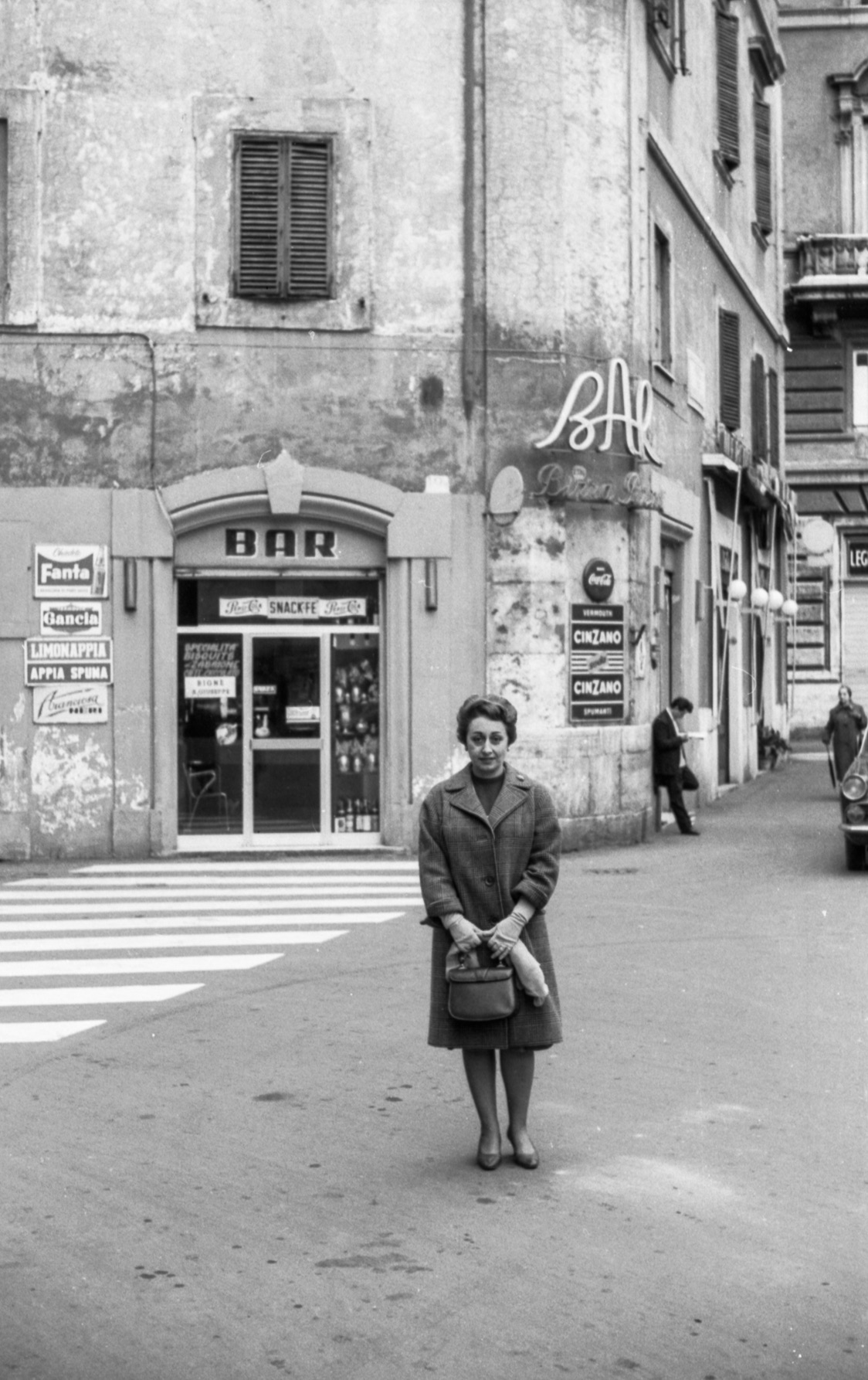 A woman in a coat stands on a city street by an old bar with vintage signs; a few people and crosswalk appear on the quiet street.