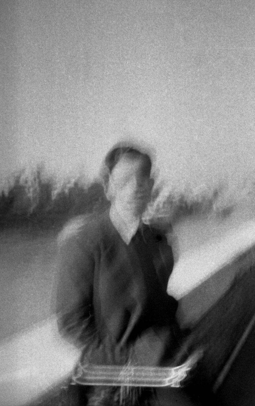 A ghostly, black and white blurry photo of a person in a coat by a river railing; out of focus with dreamlike background.
