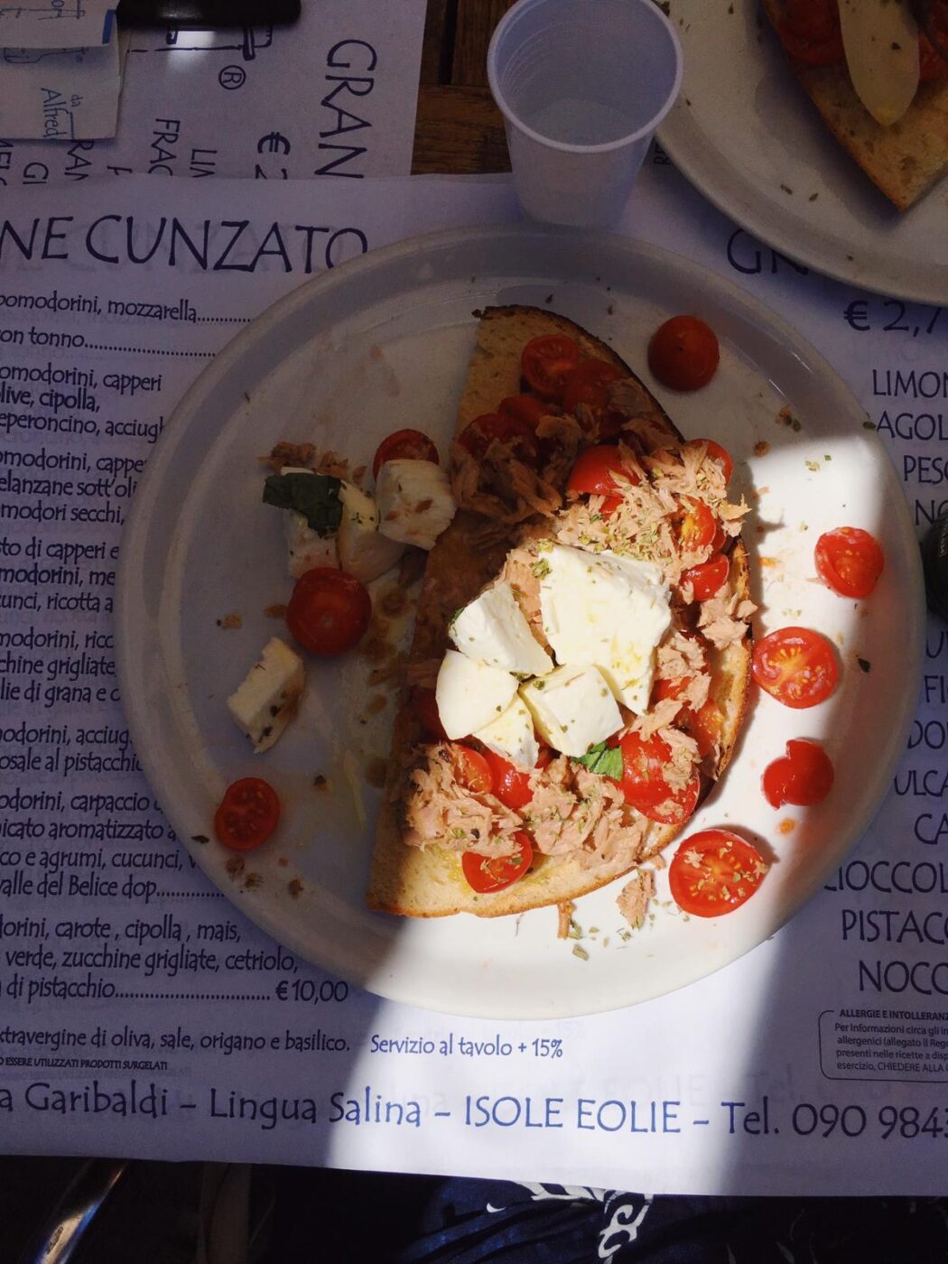 A plate with toasted bread, cherry tomatoes, mozzarella, tuna, and basil on a placemat; cup and utensils partially visible in sunlight.