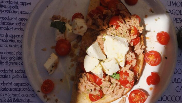 A plate with toasted bread, cherry tomatoes, mozzarella, tuna, and basil on a placemat; cup and utensils partially visible in sunlight.