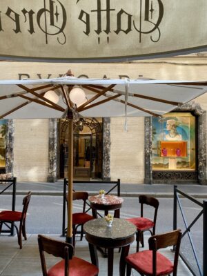 Outdoor café scene with red chairs under a white umbrella; across the street, Bulgari store window displays luxury items and a portrait.
