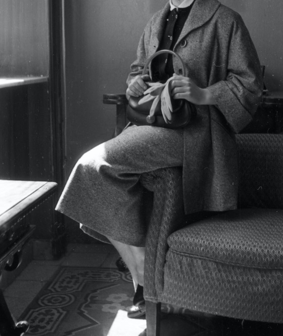 A woman in a mid-length skirt and coat perches on a chair arm, holding a handbag with gloves; her face unseen in this B&W photo.