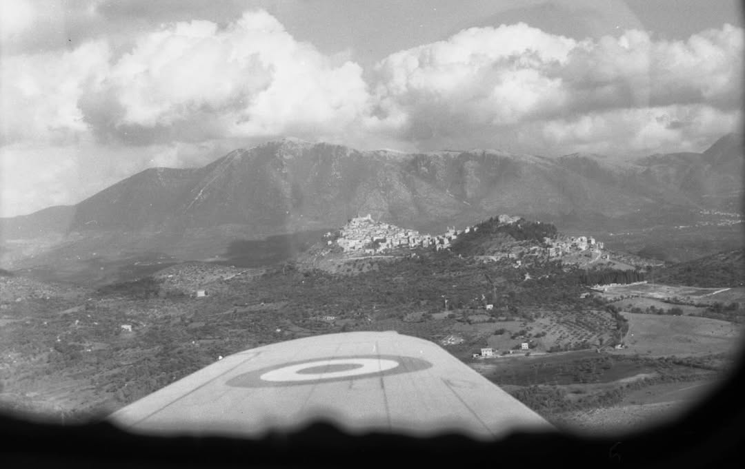 Aerial view from a plane, wing with roundel visible. Below, a small town among hills and mountains under large cloudy skies.
