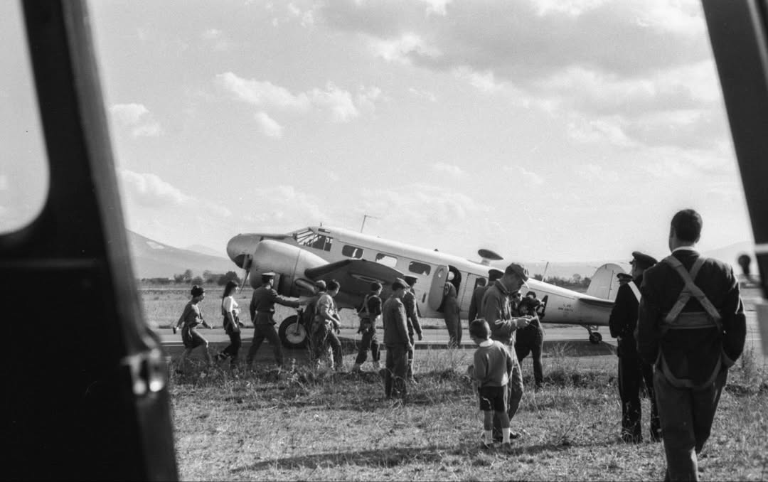 People gather by a vintage airplane on grassy airfield; some in uniform, children present. Dark objects frame scene at sides.