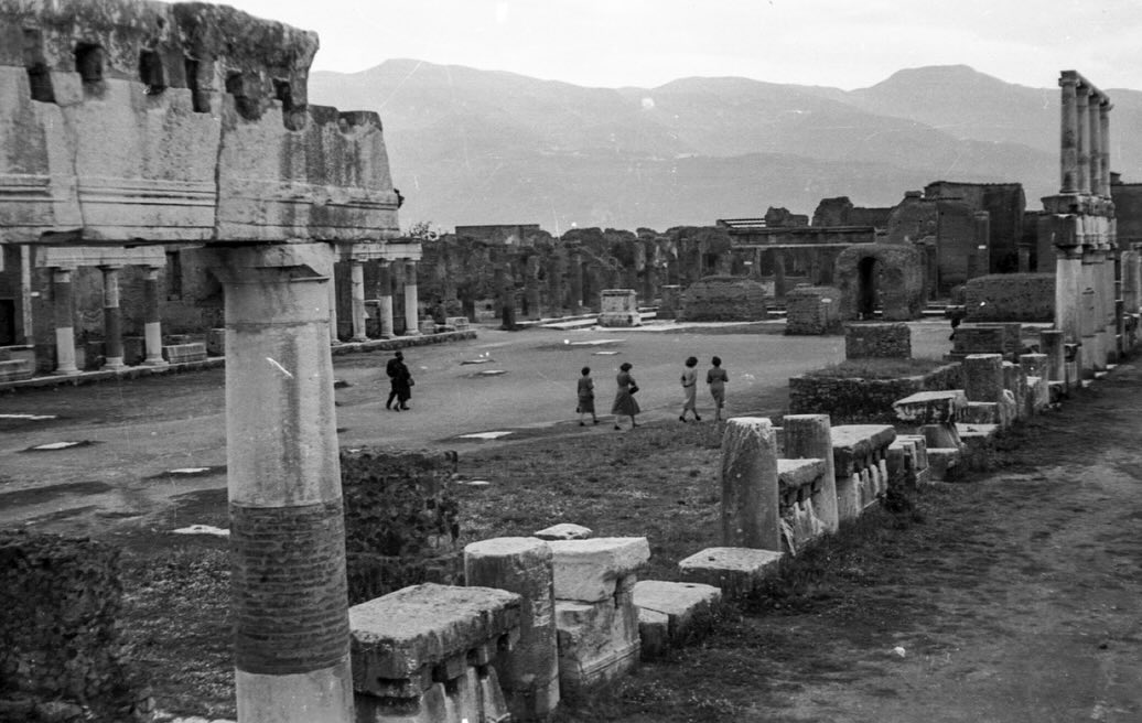 A black and white photo of Pompeii ruins: columns, stone structures, group walking on path, mountains in background.