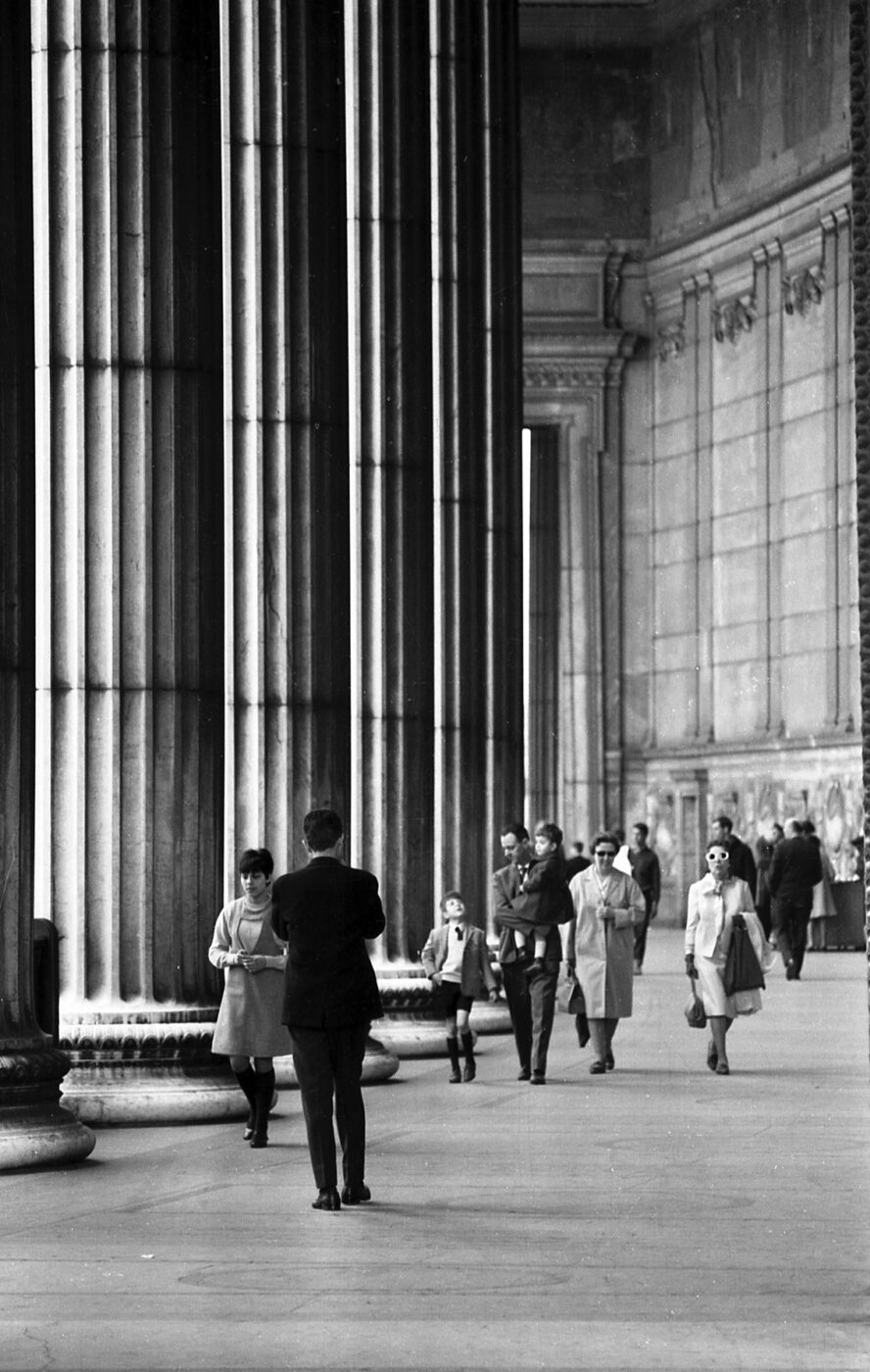 Black and white photo of people near tall columns outside a grand building, some reading or talking, in mid-20th-century attire.