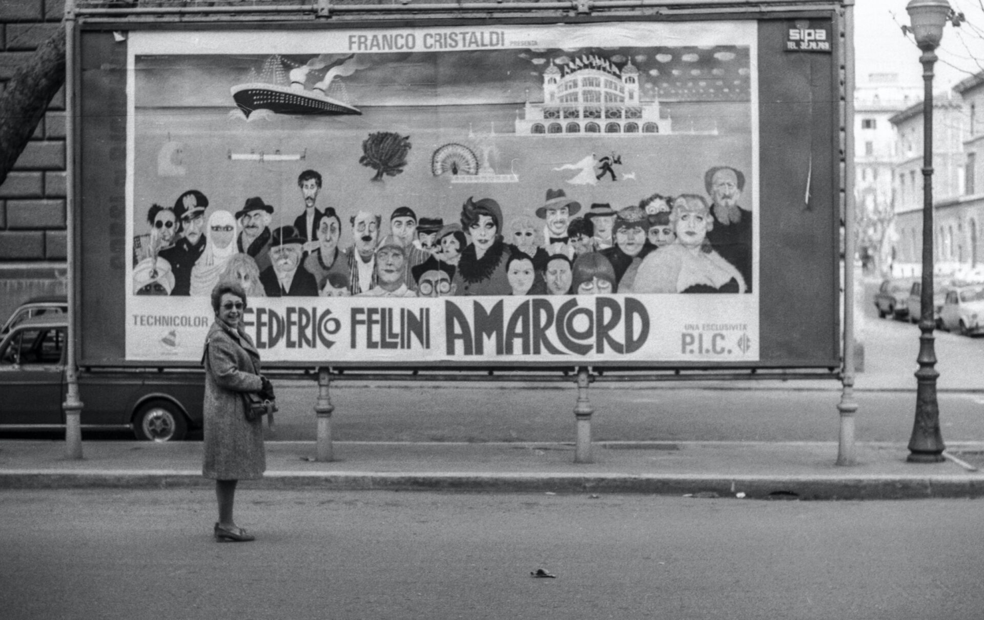 Federico Fellini Amarcord - A woman stands on a city street near a colorful billboard; vintage cars and buildings fill the background.