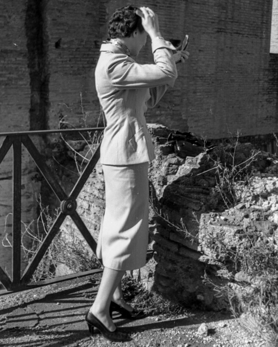 A woman in a pinstripe skirt suit and heels adjusts her hair with a compact mirror near a stone fence; weathered brick wall behind.
