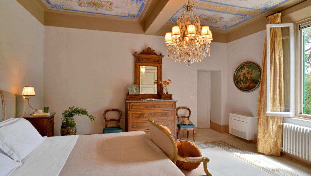 Elegant bedroom with chandelier, ornate ceiling, antique furniture, and bed by open window with gold curtains and natural light.