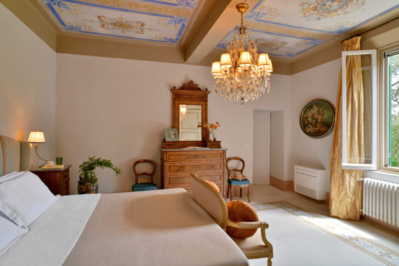 Elegant bedroom with chandelier, ornate ceiling, antique furniture, and bed by open window with gold curtains and natural light.