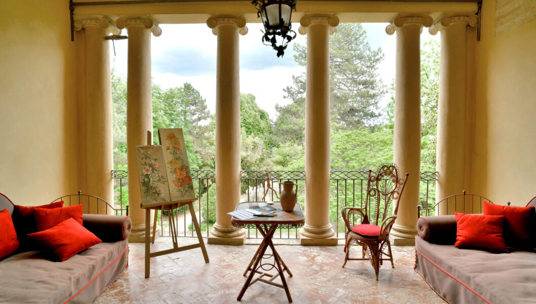 Covered veranda with four columns, red-cushioned couches, table, wicker chair, painting easel; view of a lush green garden.