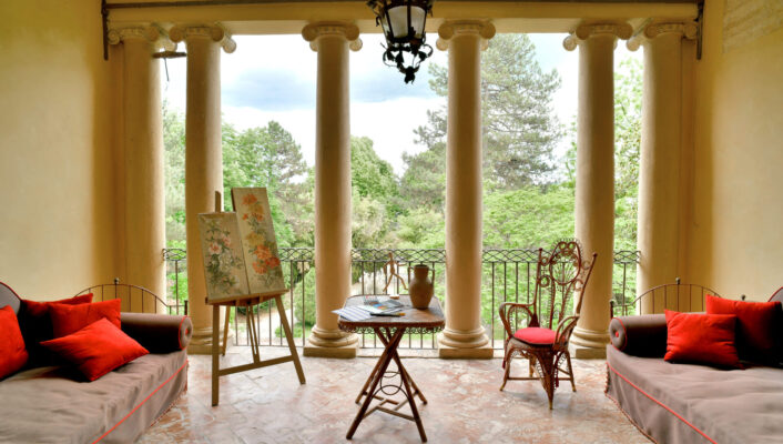 Covered veranda with four columns, red-cushioned couches, table, wicker chair, painting easel; view of a lush green garden.