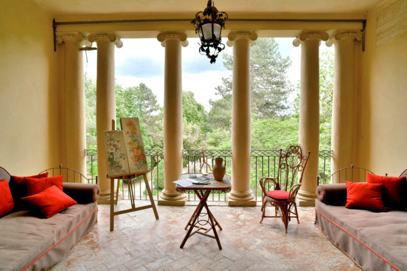 Covered veranda with four columns, red-cushioned couches, table, wicker chair, painting easel; view of a lush green garden.