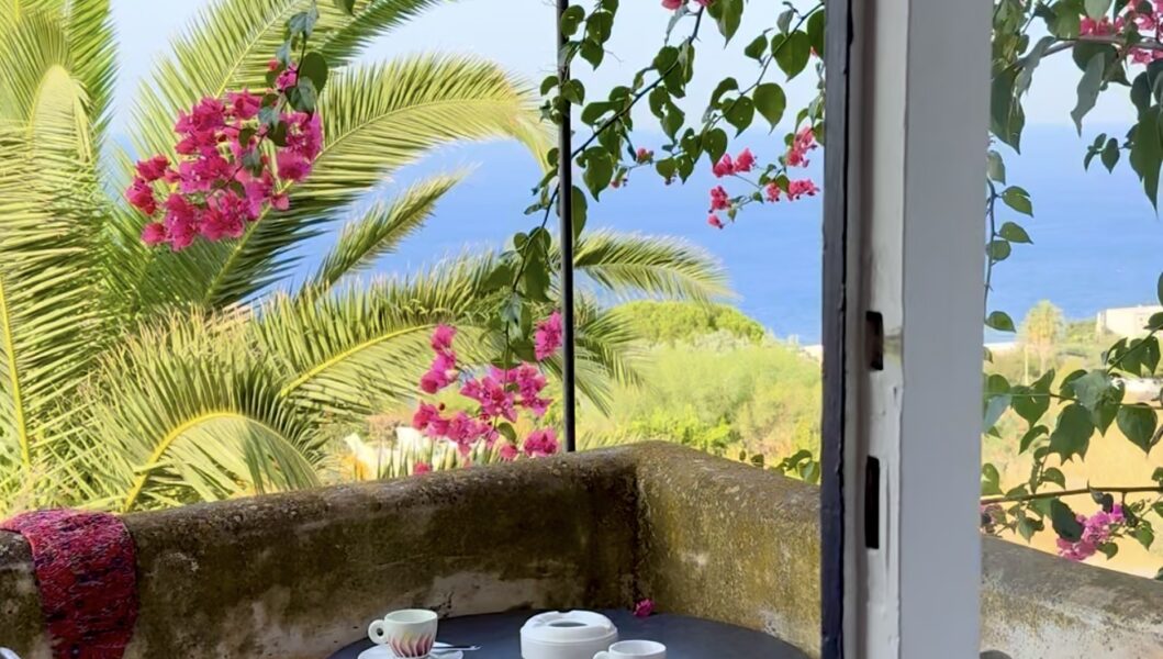 A small patio with a round table for two, cups and plates, lush palms, pink flowers, and a sea view under a clear sky.