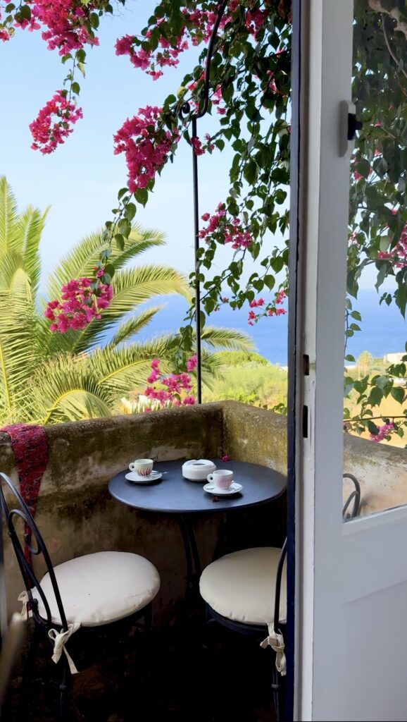 A small patio with a round table for two, cups and plates, lush palms, pink flowers, and a sea view under a clear sky.