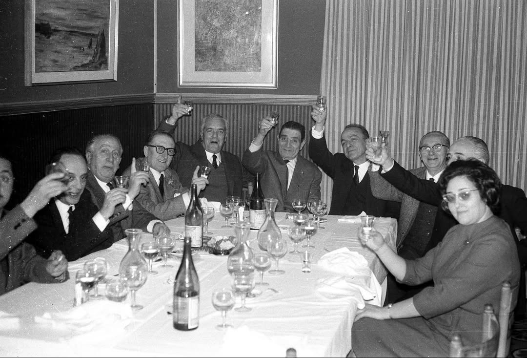 A group of people raise glasses in a toast around a formal dining table set with bottles, dishes, and glassware, celebrating together.