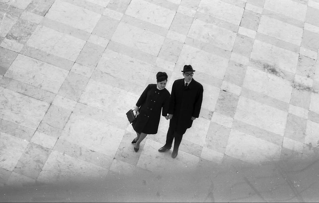 A man and woman in formal 1960s attire hold hands on a vast checkered pavement, looking up. Black-and-white, top-down view.
