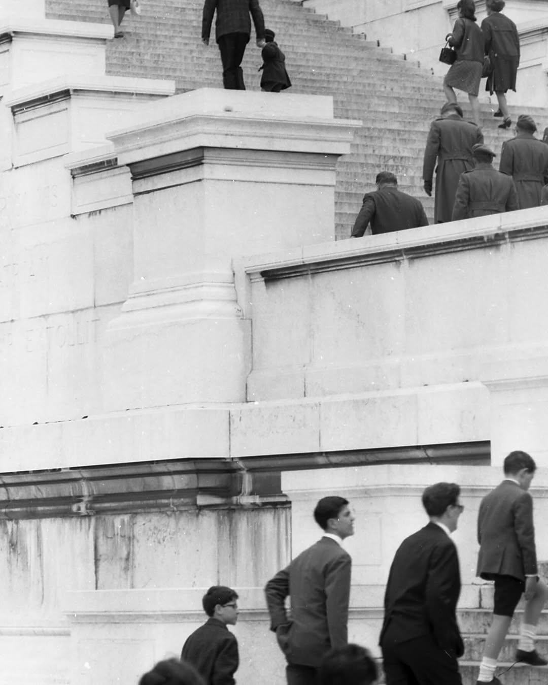 Black and white photo of adults and children in formal wear ascending stone steps of a grand building with pillars and high walls.