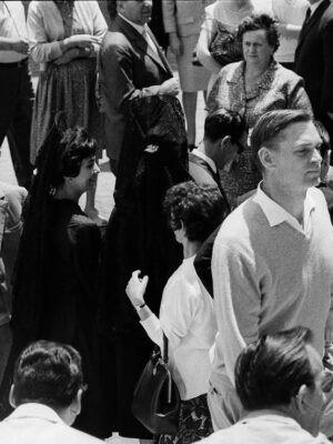 A black-and-white photo of men and women in 1960s clothes, chatting outdoors. A man in a light sweater stands front and center.