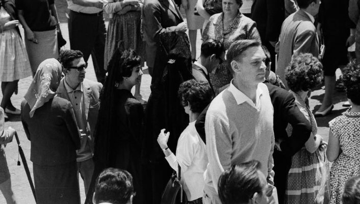 A black-and-white photo of men and women in 1960s clothes, chatting outdoors. A man in a light sweater stands front and center.