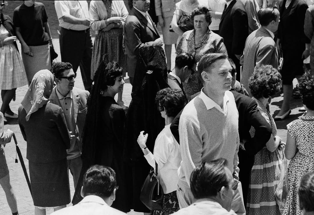 A black-and-white photo of men and women in 1960s clothes, chatting outdoors. A man in a light sweater stands front and center.